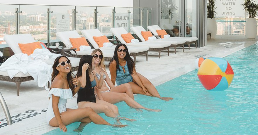 A group of girlfriends at Hotel X Toronto's rooftop pool with a beach ball, laughing.